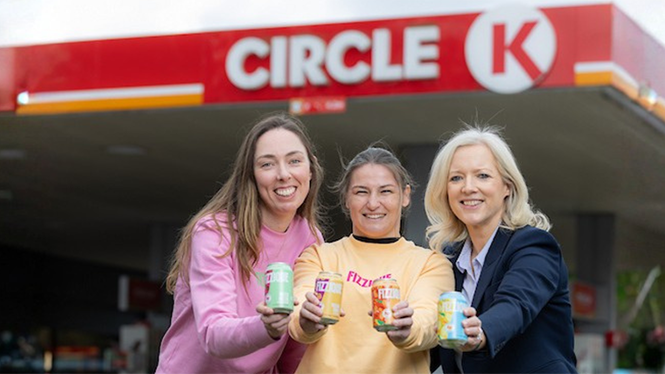 Three women holding cans of Fizzique, posing in front of a Circle K station