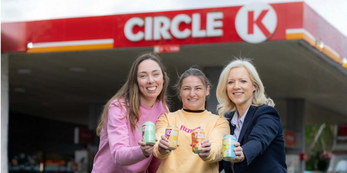 Three women holding cans of Fizzique, posing in front of a Circle K station