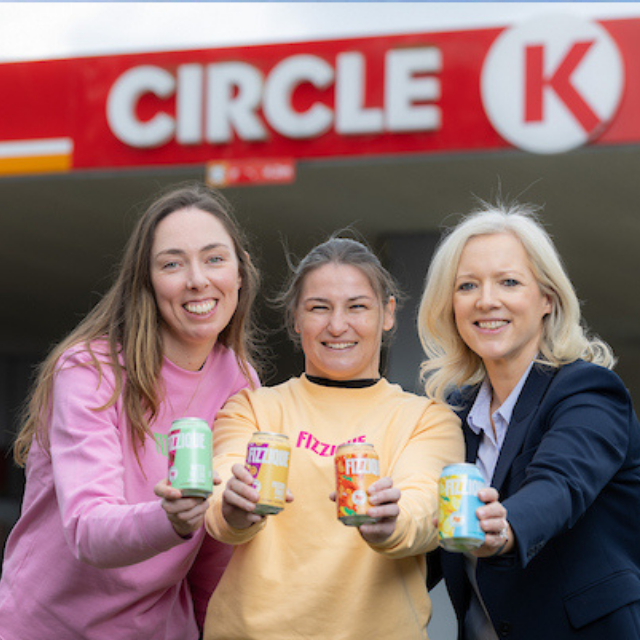 Three women holding cans of Fizzique, posing in front of a Circle K station